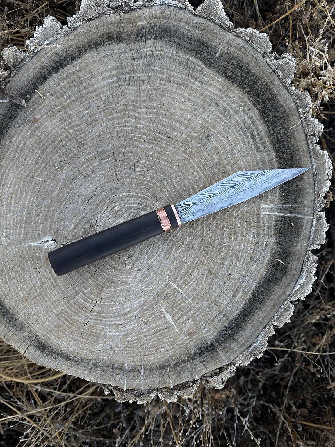 Finished broken-back seax with patterned blade, dark handle and copper fittings on a tree stump
